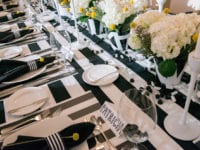 Long rectangular table set with everything in black and white stripes including table cloth, flower pots, place matts, napkin ring. Beautiful repeating centerpieces of white hydrangeas and yellow ball and silver ball shaped accent pieces, white candles in white candle stick holders.